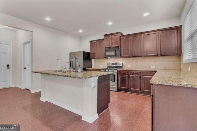 kitchen featuring dark wood-type flooring, stainless steel appliances, backsplash, a kitchen island with sink, and light stone counters
