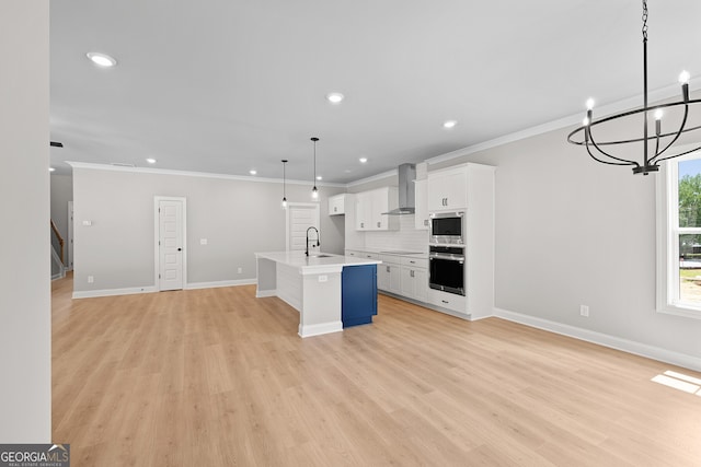 kitchen featuring appliances with stainless steel finishes, white cabinetry, decorative light fixtures, wall chimney range hood, and an island with sink