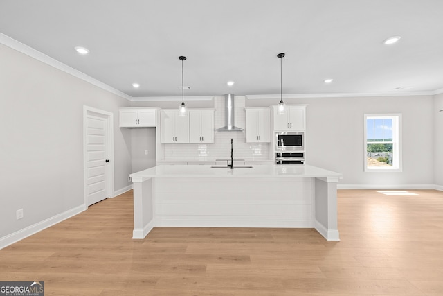 kitchen featuring a center island with sink, decorative light fixtures, appliances with stainless steel finishes, white cabinets, and wall chimney exhaust hood