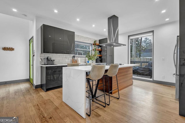 kitchen featuring gray cabinets, backsplash, a kitchen breakfast bar, a center island, and island range hood