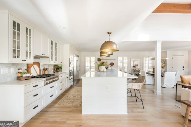 kitchen with decorative columns, white cabinetry, an island with sink, hanging light fixtures, and stainless steel appliances