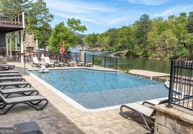 view of pool featuring an outdoor stone fireplace, fence, a water view, and a patio area