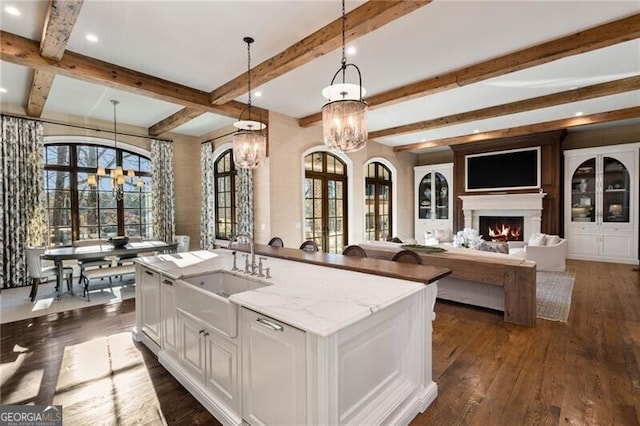 kitchen featuring an inviting chandelier, hanging light fixtures, light stone counters, an island with sink, and white cabinets