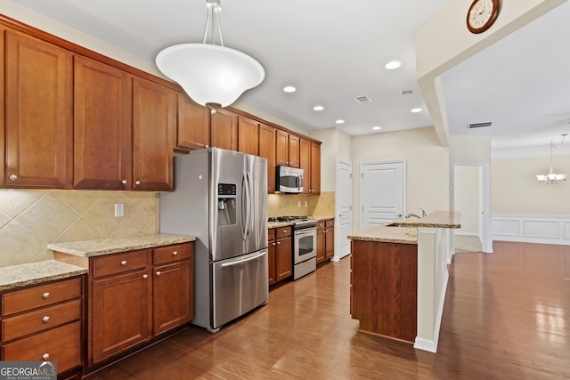 kitchen featuring wood-type flooring, appliances with stainless steel finishes, decorative light fixtures, light stone counters, and a center island with sink