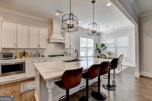 kitchen with white cabinetry, premium range hood, hanging light fixtures, and a kitchen island with sink