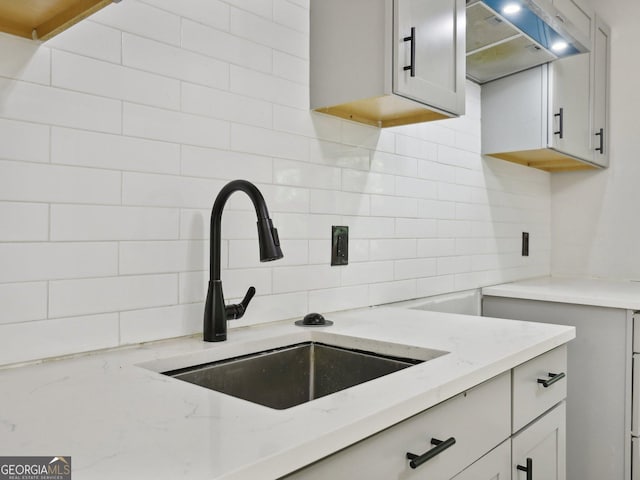 kitchen with wall chimney range hood, light stone counters, decorative backsplash, and a sink