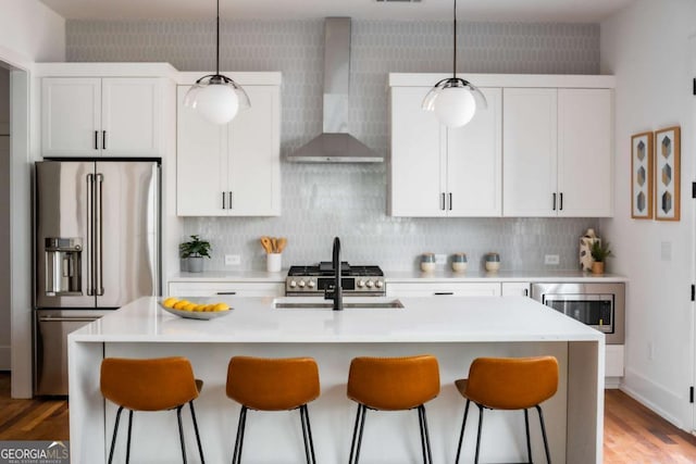 kitchen with wall chimney range hood, stainless steel appliances, white cabinetry, and a center island with sink