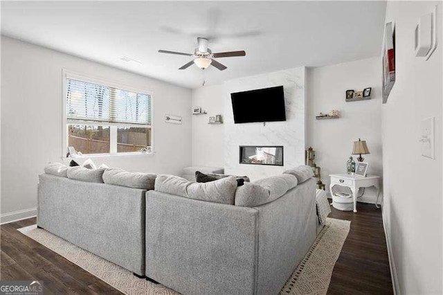 living room featuring dark wood-type flooring, a premium fireplace, a ceiling fan, and baseboards