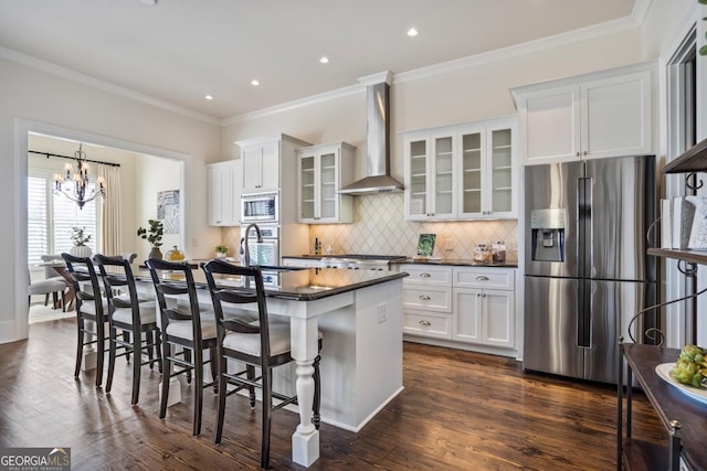 kitchen featuring wall chimney range hood, stainless steel appliances, an island with sink, white cabinets, and a kitchen bar