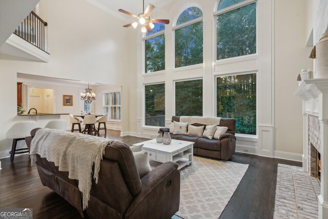 living room featuring a brick fireplace, a high ceiling, ceiling fan with notable chandelier, and dark wood-type flooring