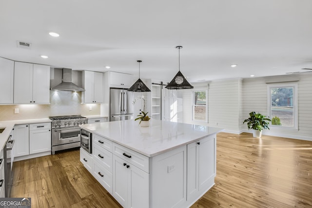 kitchen with a kitchen island, white cabinetry, premium appliances, hanging light fixtures, and wall chimney range hood