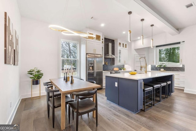 kitchen with stainless steel refrigerator with ice dispenser, backsplash, white cabinetry, wall chimney range hood, and an island with sink