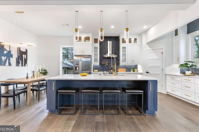 kitchen featuring stainless steel fridge with ice dispenser, white cabinetry, hanging light fixtures, wall chimney exhaust hood, and a large island with sink