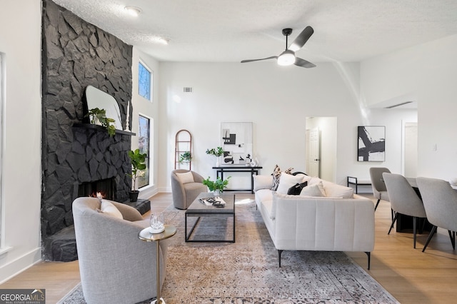 living room featuring a towering ceiling, a stone fireplace, a textured ceiling, light wood-type flooring, and ceiling fan