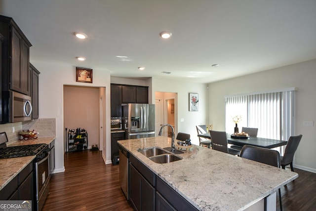 kitchen with sink, appliances with stainless steel finishes, a kitchen island with sink, dark hardwood / wood-style floors, and light stone counters