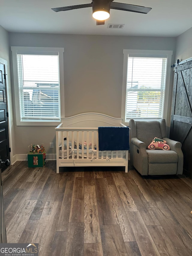 bedroom featuring dark wood-type flooring, ceiling fan, a nursery area, and a barn door