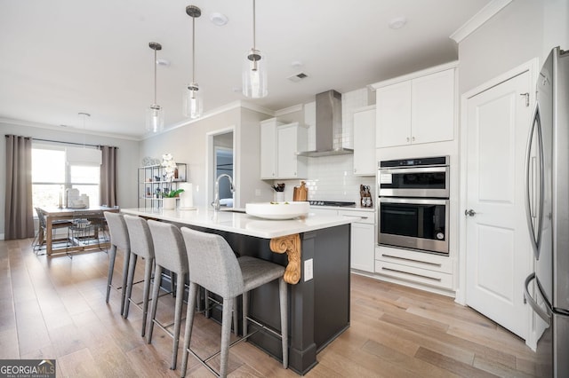 kitchen featuring white cabinetry, a center island with sink, pendant lighting, stainless steel appliances, and wall chimney range hood