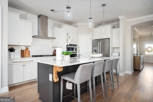 kitchen with stainless steel appliances, white cabinets, a center island with sink, decorative light fixtures, and wall chimney exhaust hood