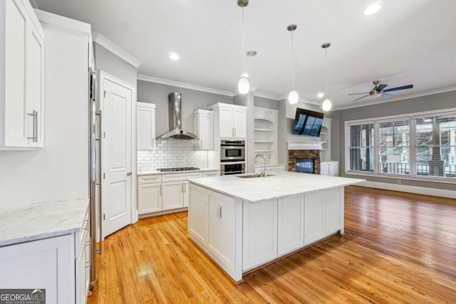 kitchen featuring wall chimney range hood, white cabinetry, backsplash, an island with sink, and decorative light fixtures