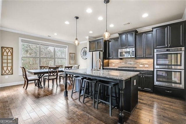 kitchen featuring appliances with stainless steel finishes, an island with sink, a kitchen bar, hanging light fixtures, and light stone countertops