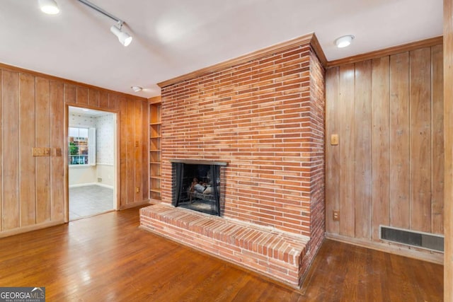 unfurnished living room featuring hardwood / wood-style flooring, rail lighting, wooden walls, ornamental molding, and a brick fireplace