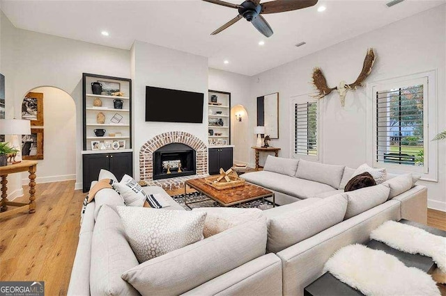 living room with built in shelves, ceiling fan, a brick fireplace, and light wood-type flooring