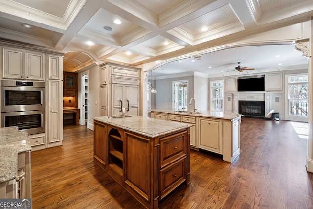 kitchen featuring stainless steel double oven, ornamental molding, light stone countertops, and an island with sink