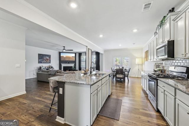 kitchen featuring sink, ornamental molding, appliances with stainless steel finishes, light stone countertops, and a kitchen island with sink