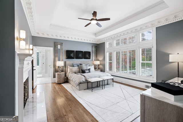 bedroom with crown molding, ceiling fan, a fireplace, a raised ceiling, and light wood-type flooring