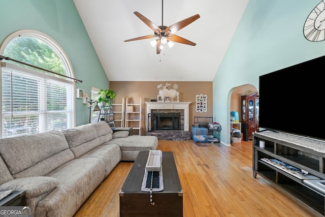 living room with ceiling fan, a stone fireplace, high vaulted ceiling, and light wood-type flooring