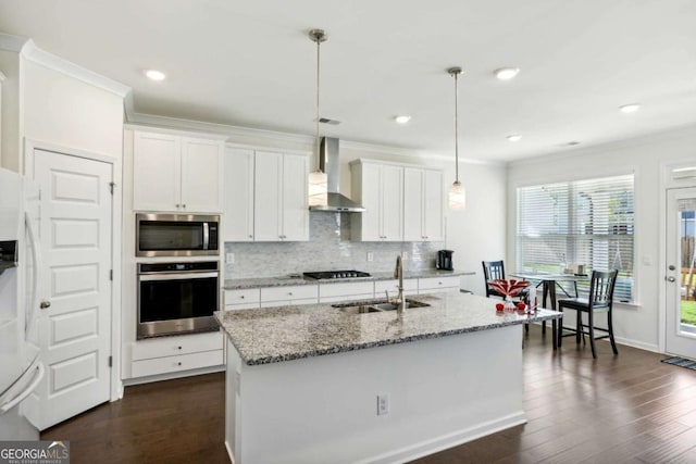 kitchen with appliances with stainless steel finishes, decorative light fixtures, an island with sink, sink, and wall chimney range hood