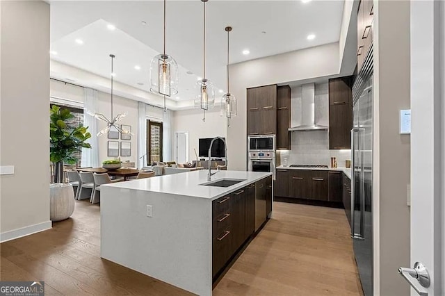 kitchen with wall chimney exhaust hood, sink, decorative light fixtures, light hardwood / wood-style flooring, and a kitchen island with sink