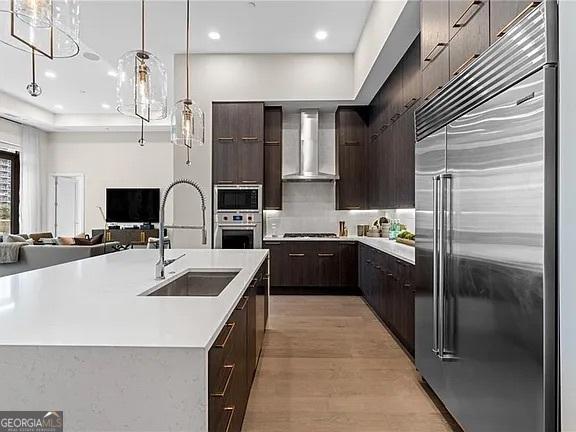 kitchen featuring wall chimney exhaust hood, sink, built in appliances, a center island with sink, and pendant lighting