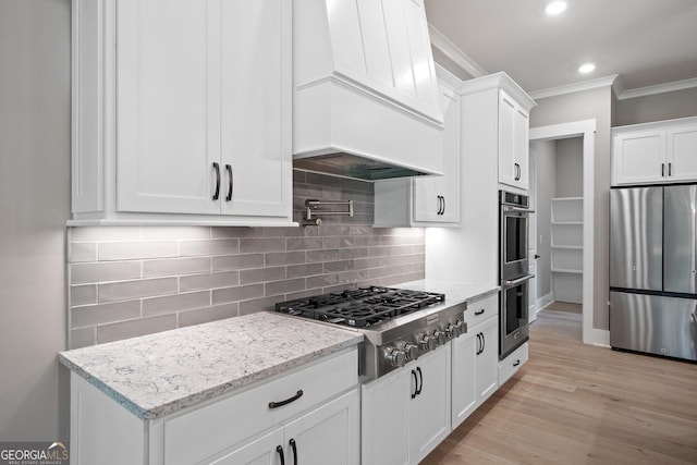 kitchen featuring appliances with stainless steel finishes, white cabinetry, crown molding, light stone countertops, and custom range hood