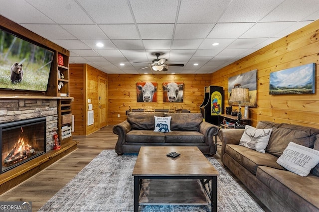 living room featuring hardwood / wood-style flooring, ceiling fan, a stone fireplace, and wooden walls