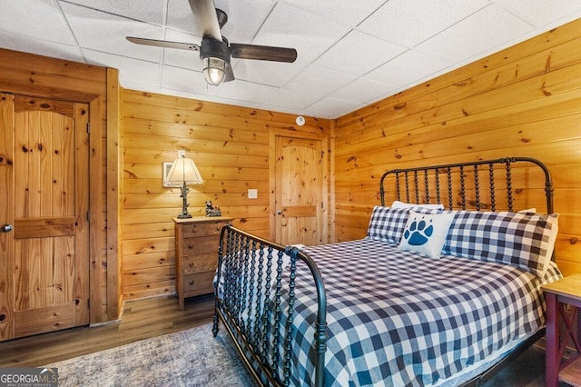 bedroom with a drop ceiling, dark hardwood / wood-style flooring, ceiling fan, and wood walls