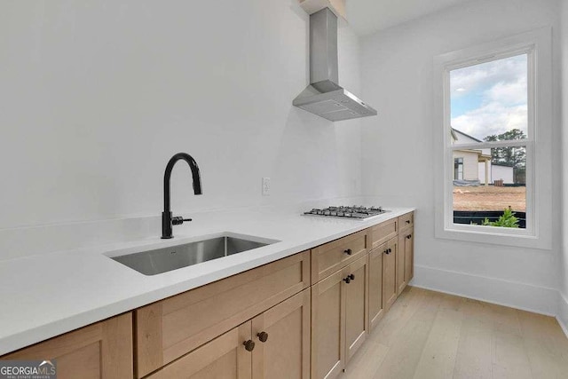 kitchen with light brown cabinetry, stainless steel gas stovetop, sink, wall chimney range hood, and light wood-type flooring