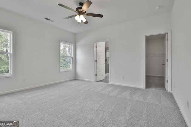 unfurnished bedroom with multiple windows, a walk in closet, light colored carpet, and ceiling fan