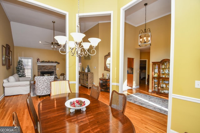 dining space with wood-type flooring, a stone fireplace, ornamental molding, and ceiling fan with notable chandelier