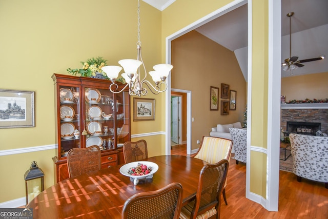 dining area featuring lofted ceiling, a stone fireplace, wood-type flooring, and ceiling fan with notable chandelier