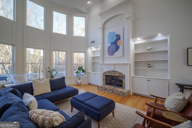 living room with a stone fireplace, a towering ceiling, light hardwood / wood-style flooring, crown molding, and built in shelves