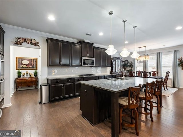 kitchen with sink, appliances with stainless steel finishes, light stone countertops, a center island with sink, and decorative light fixtures