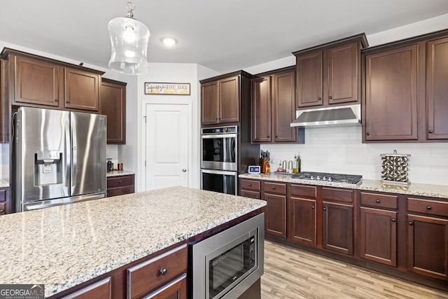 kitchen with appliances with stainless steel finishes, pendant lighting, decorative backsplash, light stone countertops, and light hardwood / wood-style flooring