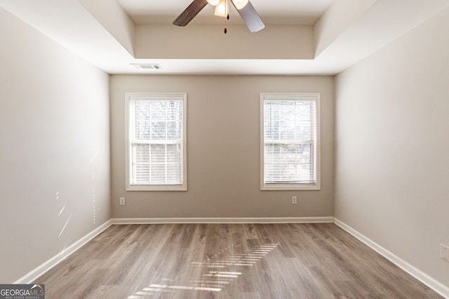 empty room with a raised ceiling, ceiling fan, a wealth of natural light, and light hardwood / wood-style floors