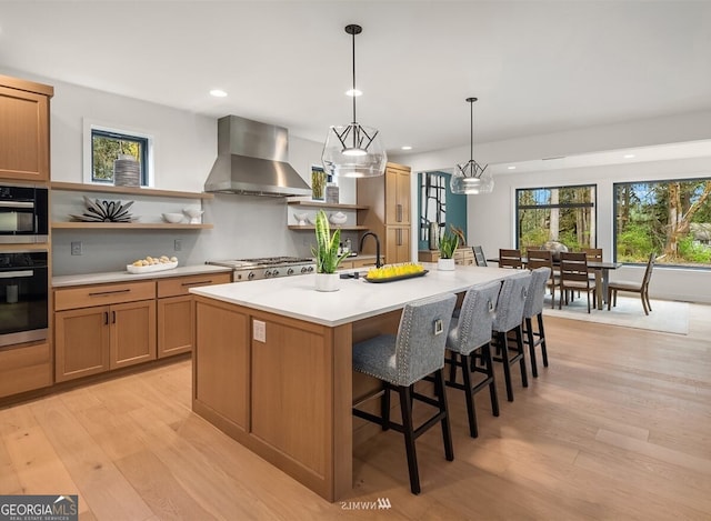 kitchen with decorative light fixtures, an island with sink, exhaust hood, light hardwood / wood-style floors, and stainless steel appliances