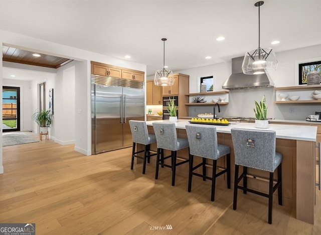 kitchen featuring a breakfast bar, range hood, hanging light fixtures, stainless steel built in fridge, and light wood-type flooring