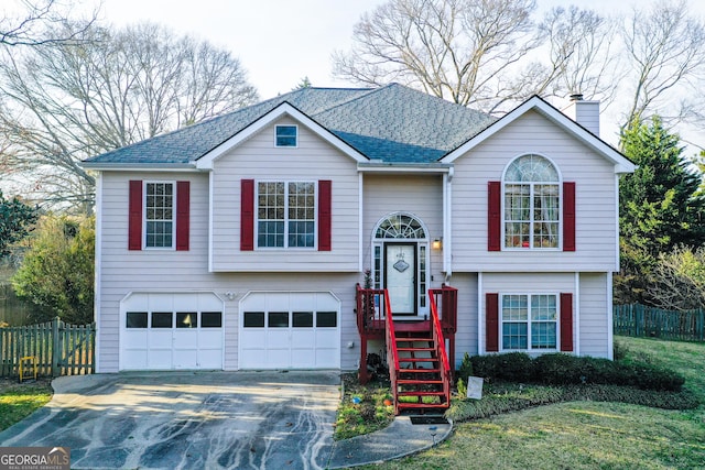 raised ranch with a chimney, driveway, an attached garage, roof with shingles, and fence