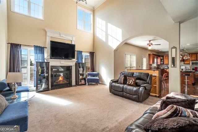 living area featuring arched walkways, a ceiling fan, a fireplace with flush hearth, ornamental molding, and carpet