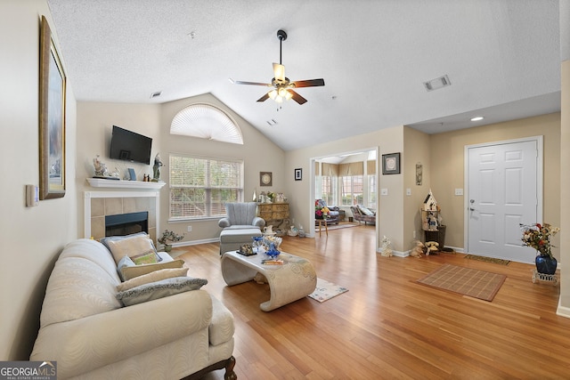 living room featuring lofted ceiling, light hardwood / wood-style flooring, a tile fireplace, ceiling fan, and a textured ceiling