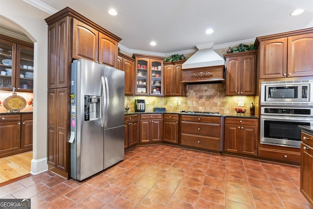 kitchen featuring premium range hood, dark stone countertops, ornamental molding, appliances with stainless steel finishes, and decorative backsplash